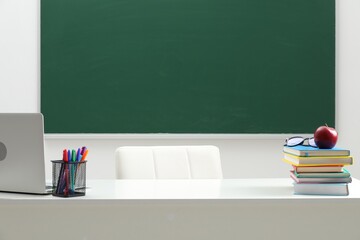 Different school stationery, laptop, glasses and red apple on white table near blackboard indoors