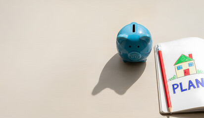 Blue piggy bank with notebook drawing of a house and the word “PLAN”, symbolizing saving money for home, financial planning, and future investment. Copy space on bright background.