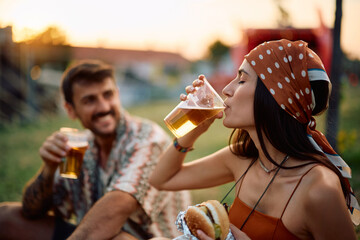 Young woman enjoying in glass of beer while eating with her boyfriend at music festival.