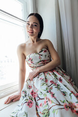 Fashionable woman in summer dress with flowers sitting by window portrait