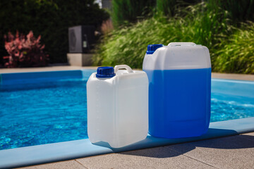 White and blue plastic chemical canisters placed on the poolside edge next to a clean blue swimming pool, used for water treatment and pool maintenance.