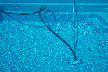 Blue hose and telescopic pole connected to a manual pool vacuum cleaner removing debris from the pool floor during outdoor summer maintenance.