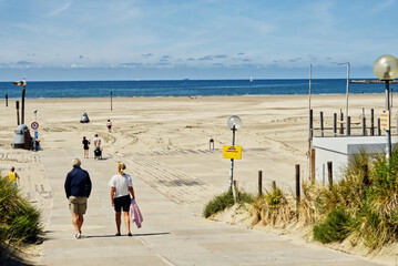Senior couple walking down paved path to wide sandy beach on sunny summer day at north sea coast in...