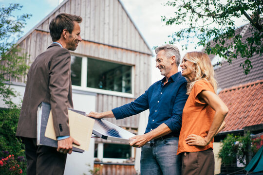 Happy real estate agent with couple holding solar panel in front of house