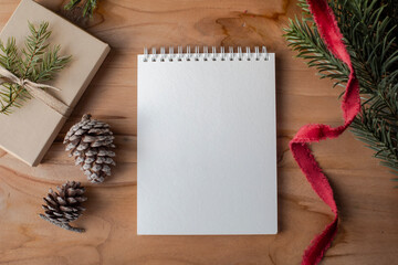 Flat lay of a blank spiral notepad on wooden table with pinecones, red ribbon, evergreen sprigs, and a wrapped gift. Perfect for holiday lists, notes, or festive messages.