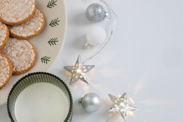 Top-down view of holiday cookies and milk prepared for Santa, with star-shaped lights and Christmas ornaments on a white background. Cozy and festive winter holidays mood.