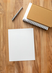 Top-down view of a blank white sheet of paper with spiral notebooks, a mustard-colored journal, and a silver pen on a wooden desk. Ideal for workspace, journaling, or creative mockups.