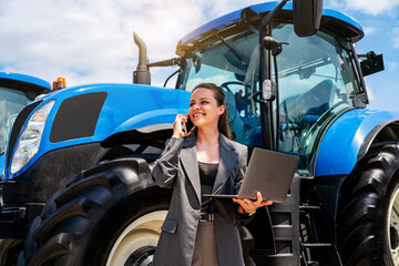 Female agricultural equipment dealer using laptop and talking on phone near farm tractor