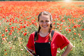 Joyful young woman ecological farmer with tablet enjoying summer day in blooming red poppy flower field