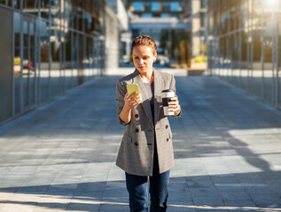 Busy modern woman texting on smartphone while holding coffee mug outdoors in city center.