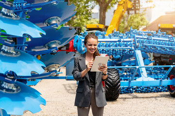 Young woman in business suit working on digital tablet near farm implements in farm equipment dealership