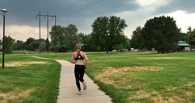 Slow motion running woman runs during stormy skies along a park pathway. Sleek and fit she inspires others to get out and run despite challenges including weather.