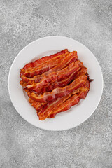 Cooked bacon for breakfast on a white plate on a gray concrete background. Top view, copy space.