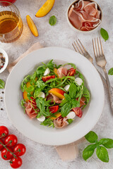 Salad with peaches, arugula, prosciutto, mozzarella cheese and cherry tomatoes with balsamic vinegar in a ceramic plate on a concrete background. Selective focus.