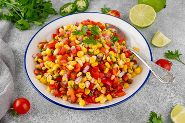 Corn salsa in a bowl with lime, jalapeno pepper, fresh herbs and tortilla chips on a concrete background. Mexican cuisine. Healthy food.