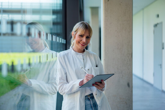 Smiling female doctor with tablet PC and digitized pen leaning on glass window in hospital