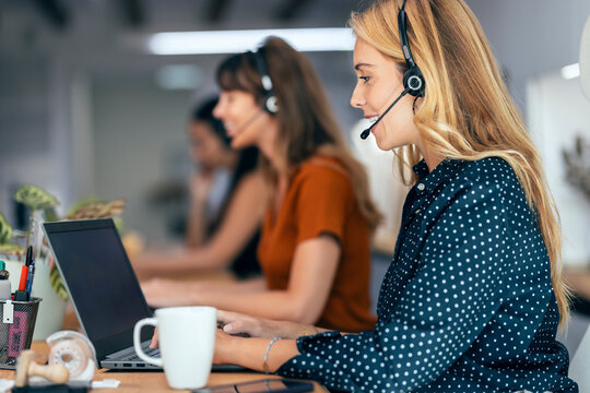 Smiling blond businesswoman talking through headset sitting by colleagues at desk in office