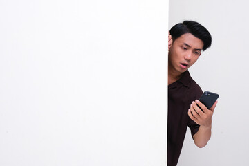 Adult Asian man peeking behind wall showing shocked expression when looking at his mobile phone