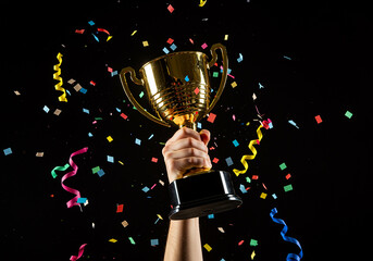 A person's hand raising a gleaming gold trophy cup in celebration, with colorful confetti showering down against a dark background.