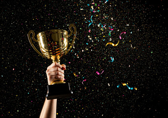 Triumphant moment of victory as a champion's hand raises a gleaming gold trophy cup amidst a shower of colorful confetti.