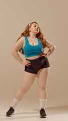 Self-assured woman in fitness clothing standing with hands on hips against beige background....