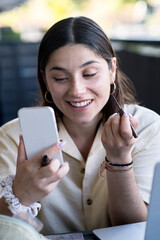 Young Woman Applying Mascara While Looking in Her Phone at a Cafe