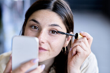 Young Woman Applying Mascara While Looking in Her Phone at a Cafe