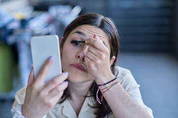 Young Woman Applying Mascara While Looking in Her Phone at a Cafe