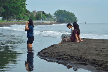 Three girls sit and talk near the sandy edge of a stream while one stands in the shallow water,...