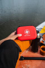 A person is sitting comfortably in a kayak, securely holding a bright red first aid kit, which is...