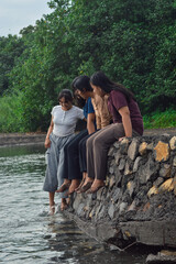 Three girls watch closely as one of them carefully steps into the cool stream near the beach, surrounded by sand and trees.