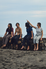 One teen girl points at something in the distance while the others watch, seated on the beach enjoying the calm morning by the sea together.