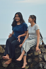 Two teenage girls sit on a beach rock barrier, chatting and enjoying the peaceful beach atmosphere in the soft morning light.