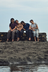 Sitting along the brackish beach edge, four girls relax close together, three resting heads on friends' shoulders while one sits upright enjoying the morning breeze.