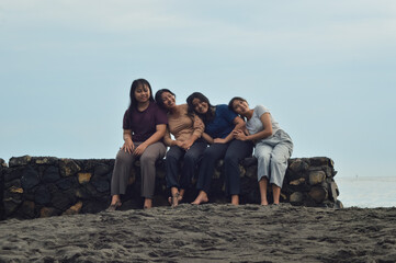 Four teen girls sit in a row along the beach, three resting on each other’s shoulders while one sits upright facing the calm sea and morning light.
