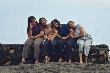 The girls share a cheerful moment, three resting heads on their friends’ shoulders while smiling, seated along the peaceful sandy shore in the early morning.