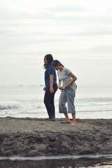 On the sandy shore, two teen girls play in the gentle morning light, one glancing down at the sand as the other smiles brightly.