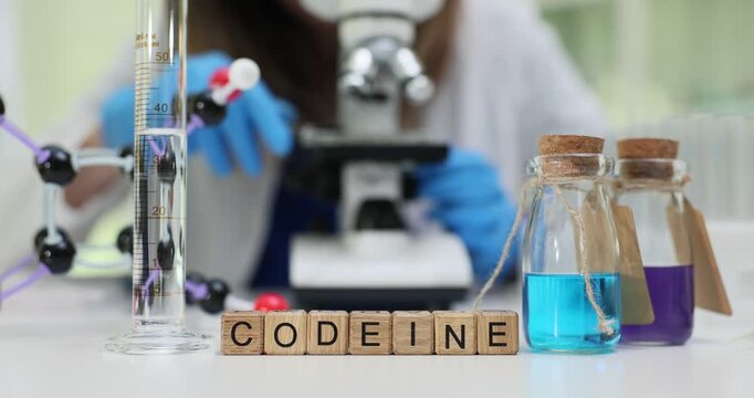 Wooden cubes show Codeine word placed on laboratory table near colorful liquids. Lab assistant checks reaction using microscope and modern equipment