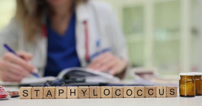 Wooden cubes show Staphylococcus placed by medical supplies and clipboard. Woman in coat examines documents controlling bacterial problem research