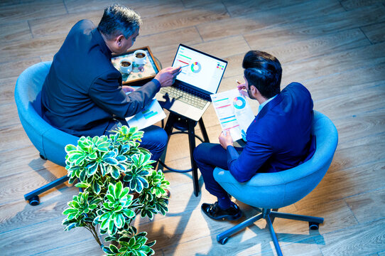 Indian corporate partners collaborate while seated at desk using laptop in top-angle office view