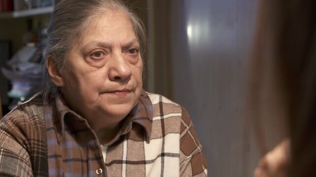 elderly woman with grey hair, wearing a plaid shirt, sits in her living room and speaks with a serious expression. Her furrowed brow and slightly pursed lips suggest she is discussing a matter of impo