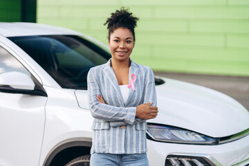 Smiling woman with pink ribbon holding arms crossed posing against her car