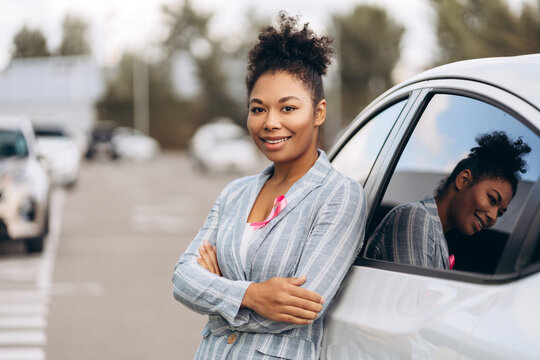 Happy woman with arms crossed wearing pink ribbon for breast cancer awareness near car