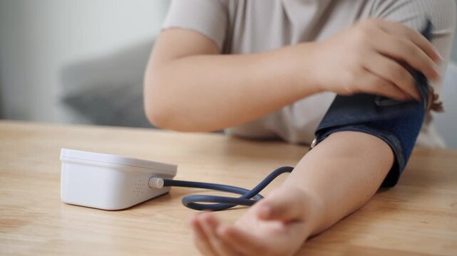 An Asian woman uses a portable blood pressure monitor in her living room. Health awareness concept.
