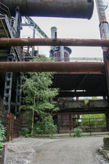 Naklejka premium Tall, rusty pipes and metal structures of a large abandoned industrial complex with trees and foliage growing through it.