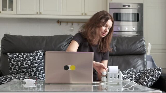 A woman refills a label printer while working on her laptop at a glass table in her modern home.
