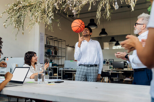 Playful businessman spinning basketball on finger by colleagues cheering in office