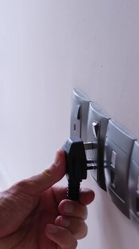 Hand plugging a black electrical plug into a wall socket. Several sockets are aligned on a white wall in a modern interior. Save energy concept.