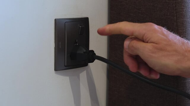 Hand pushing button on black electrical cord into one of several sockets on a white wall. Close view with focus on safe energy use. Energy use, household electricity, safe connection concept.