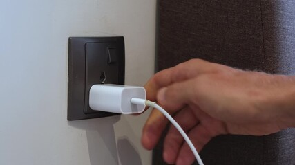 Hand inserting a white charging adapter into a wall socket. Close-up view inside a modern interior.